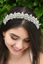 Smiling Bride wears a low tiara with crystals in pale gold. She has dark hair and green leaves behind her.