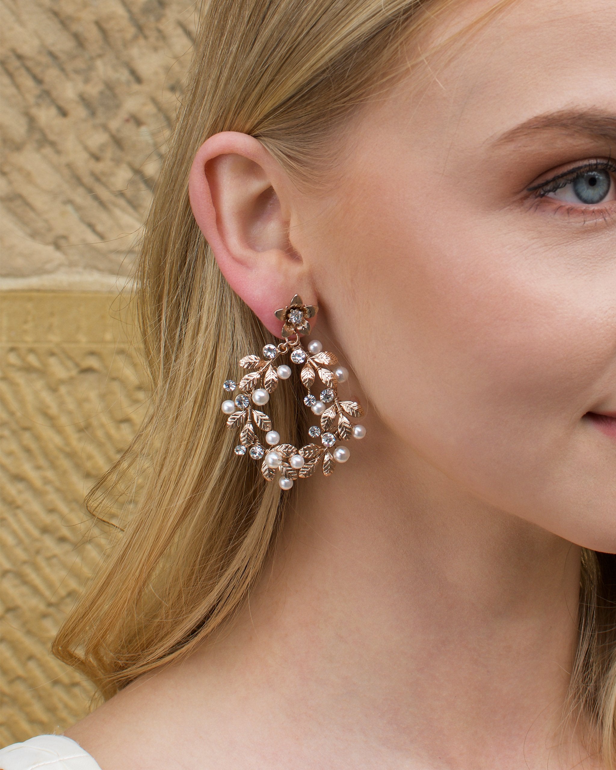 Model with blonde hair wears a rose gold hoop earring with pearls with a stone wall backdrop