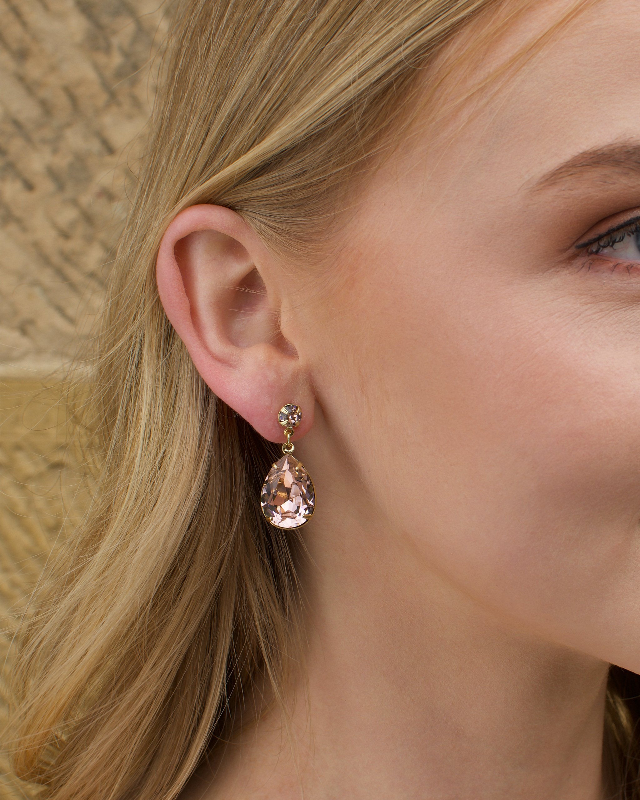 A blonde model wears a gold earring with a vintage rose colour stone with a sandstone wall background