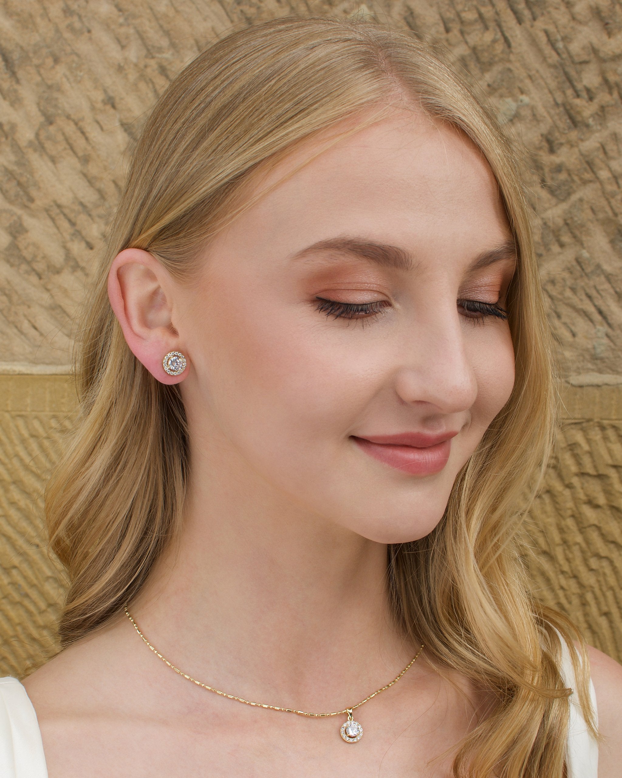 A blonde hair model wears a gold stud earring with a simple gold chain with a stone wall backdrop