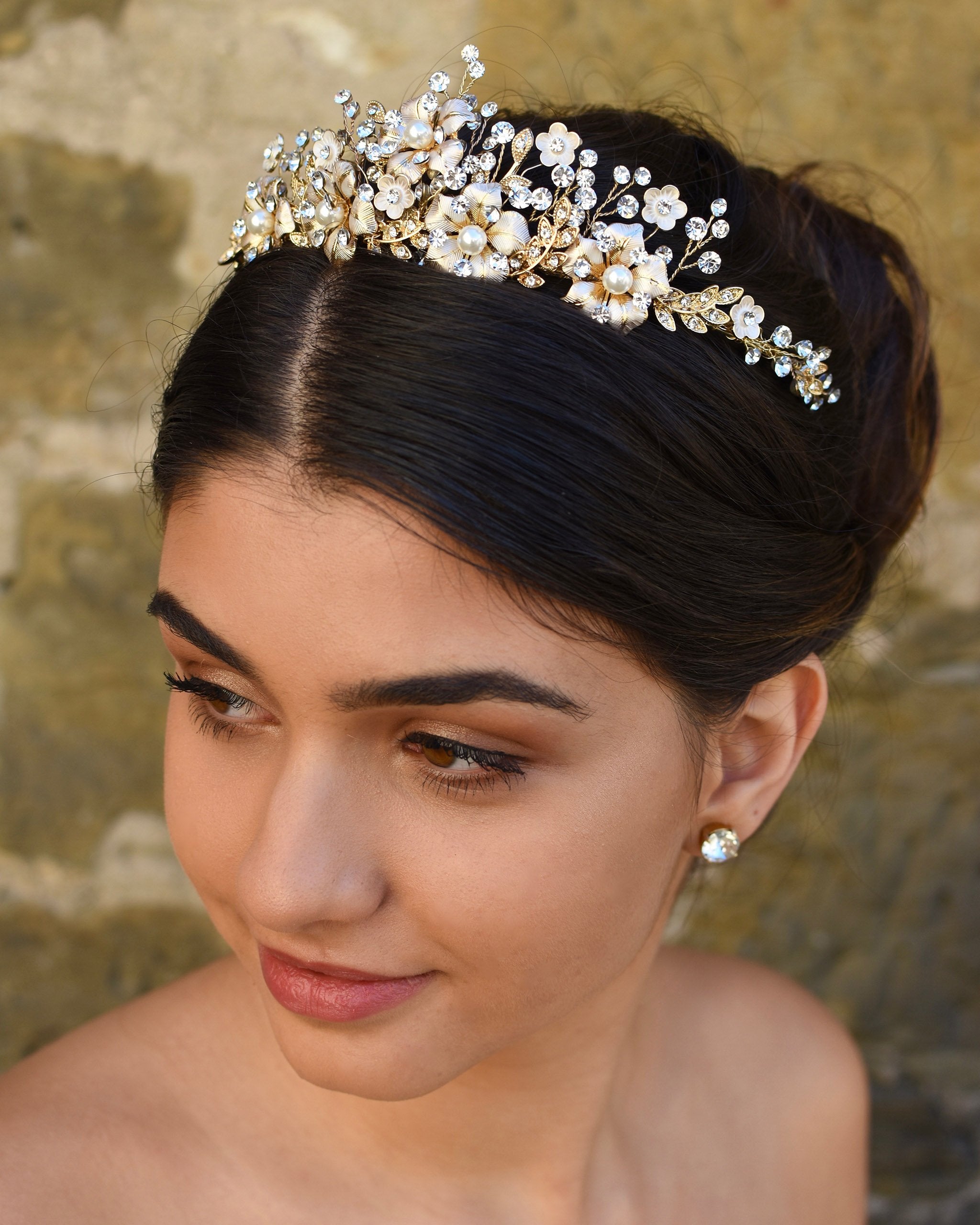 A dark haired model wears a gold tiara with pearl flowers against a stone wall background.