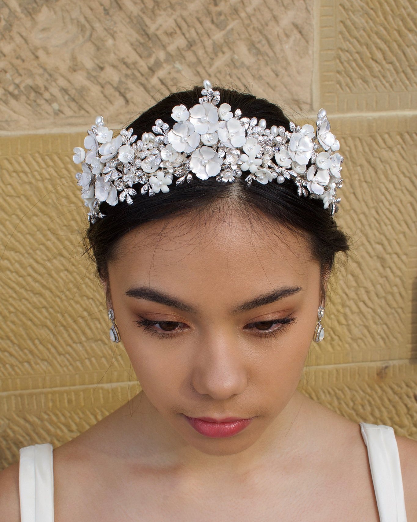 A Black haired bride wears a three pointed silver flower tiara at the front of her head. Behind is a sandstone wall.