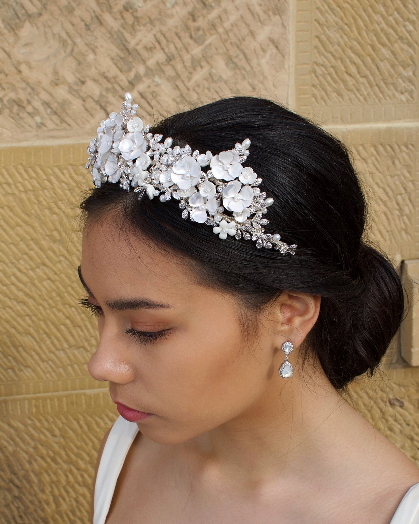 A side view of a dark haired bride wears a three pointed flower tiara at the front of her head. Behind is a sandstone wall.
