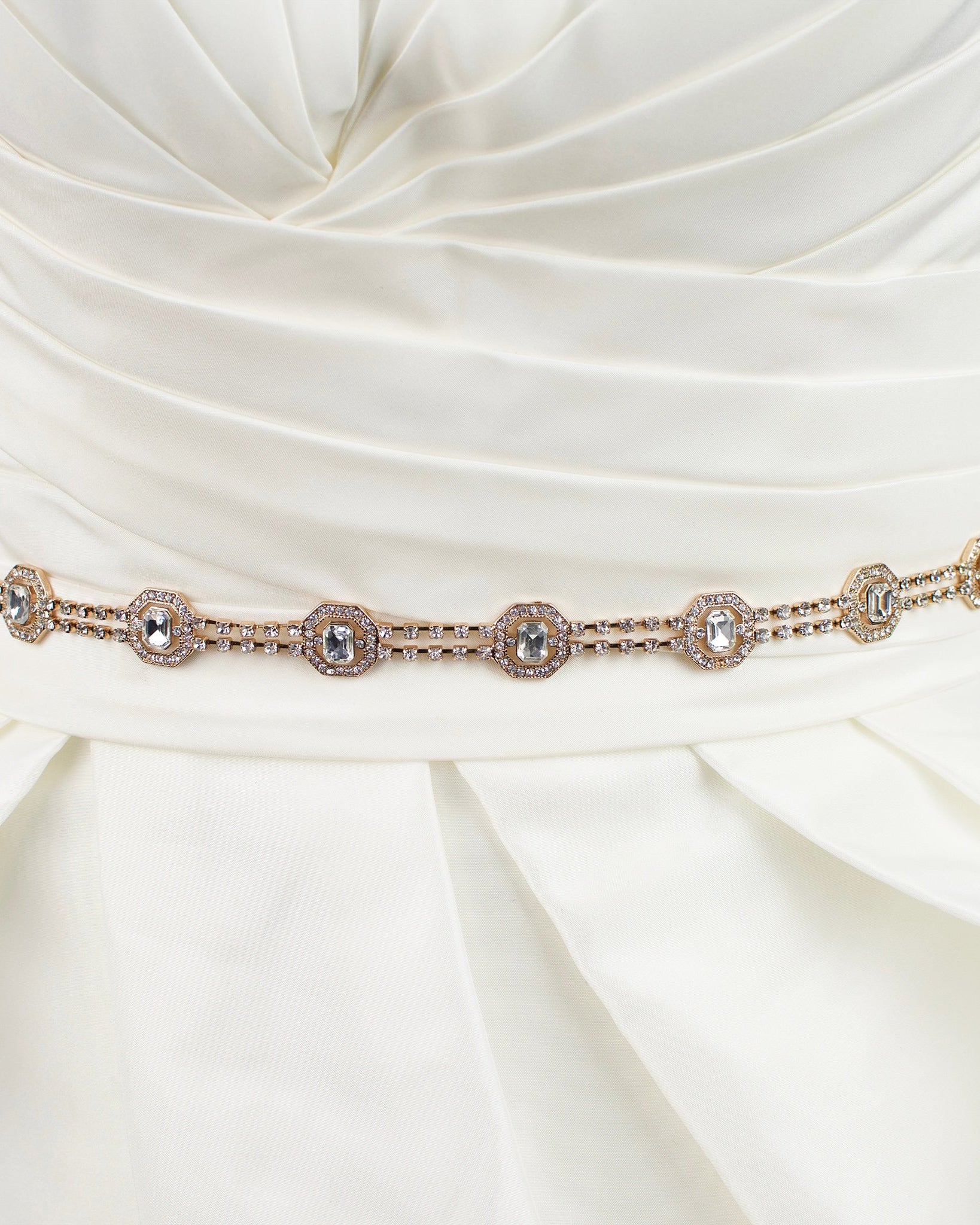 A narrow rose gold belt with crystals is fitted around the waist of an ivory bridal gown with a white background