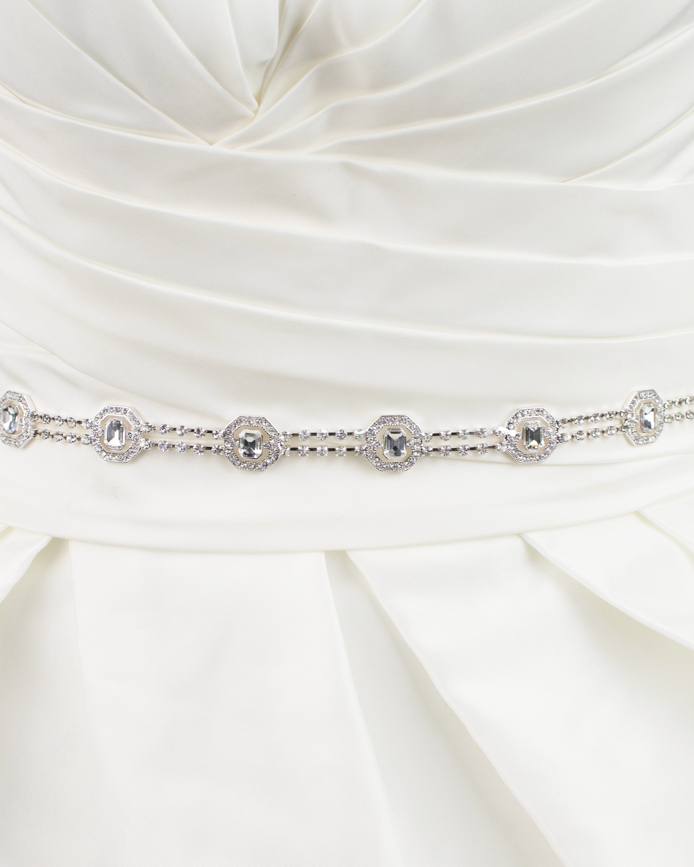 A narrow silver chain bridal belt is fitted around the waist of an ivory bridal gown with a white background