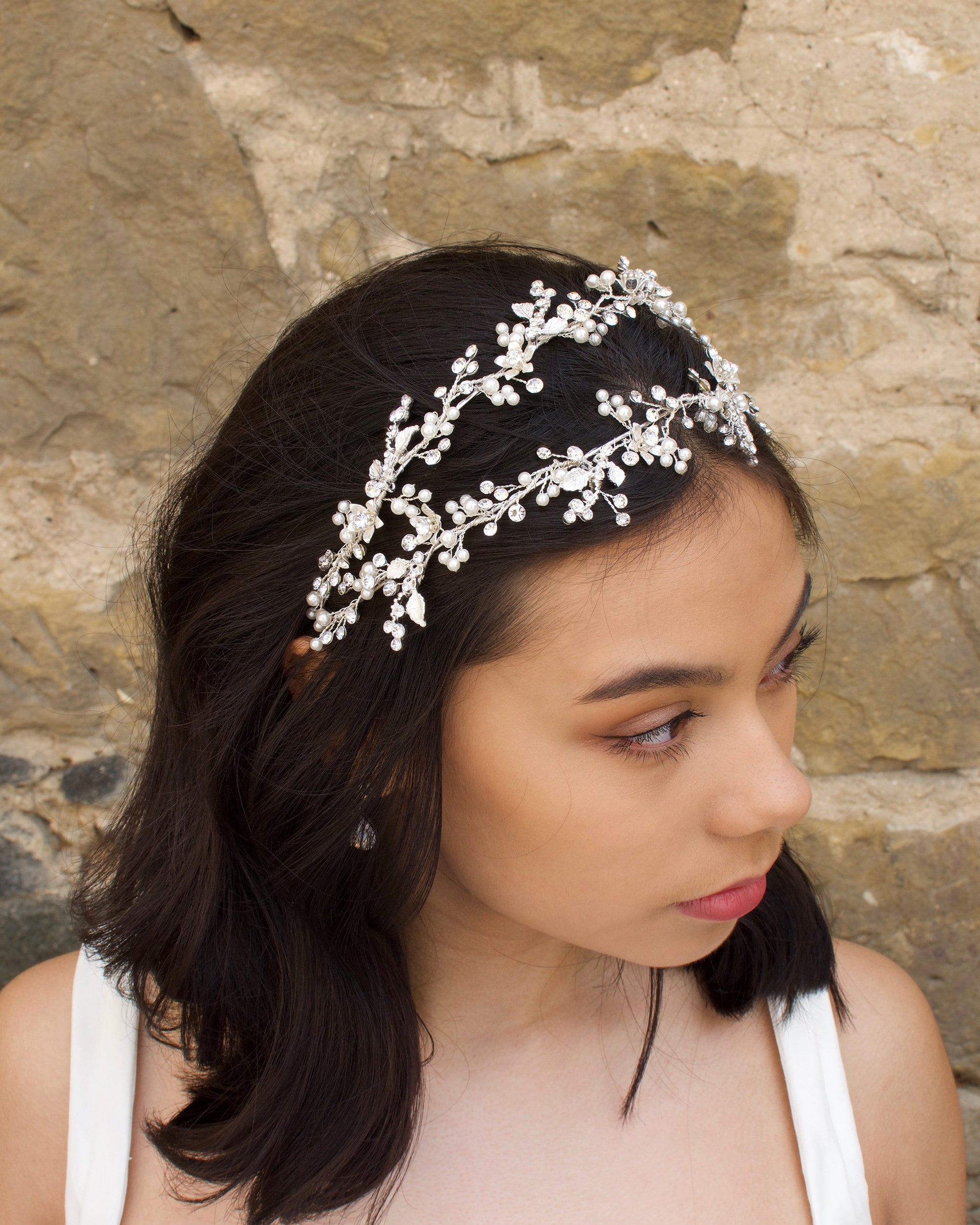 Black hair model looks to the side wearing a silver pearl handmade headband with a stone wall behind