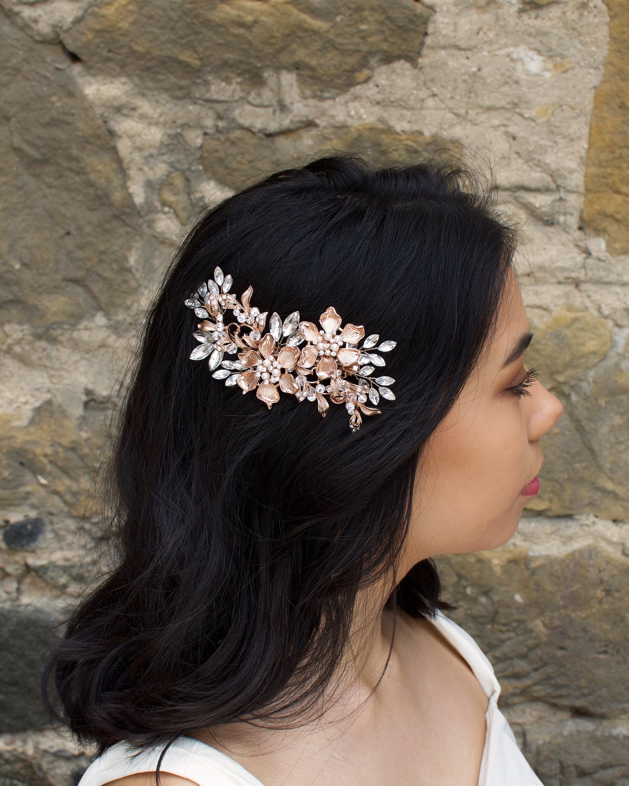 A dark haired model wears a Rose Gold flower comb on the side of her head. A stone wall is the background