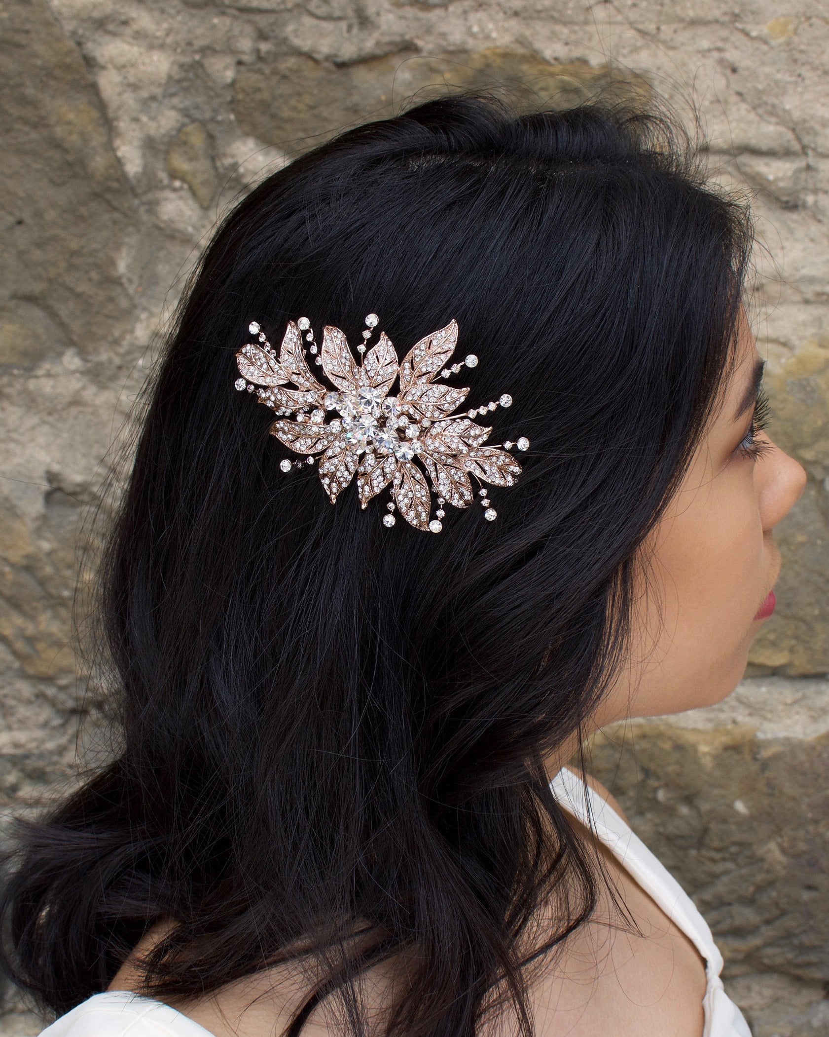 A back haired model wears a rose gold flower bridal side comb on the side of her head. A stone wall is the backdrop