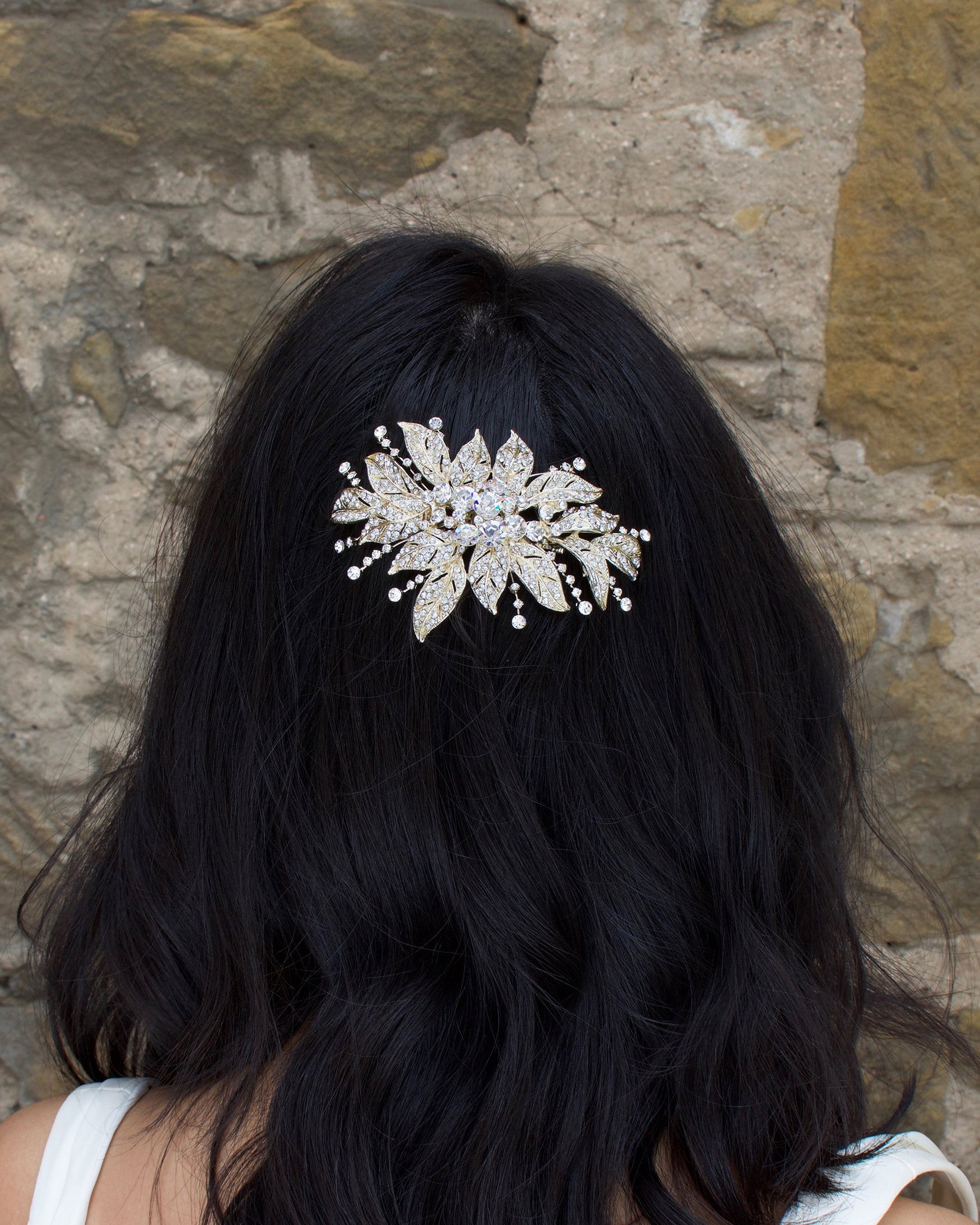 A back haired model wears a gold flower bridal side comb on the back of her head. A stone wall is the backdrop