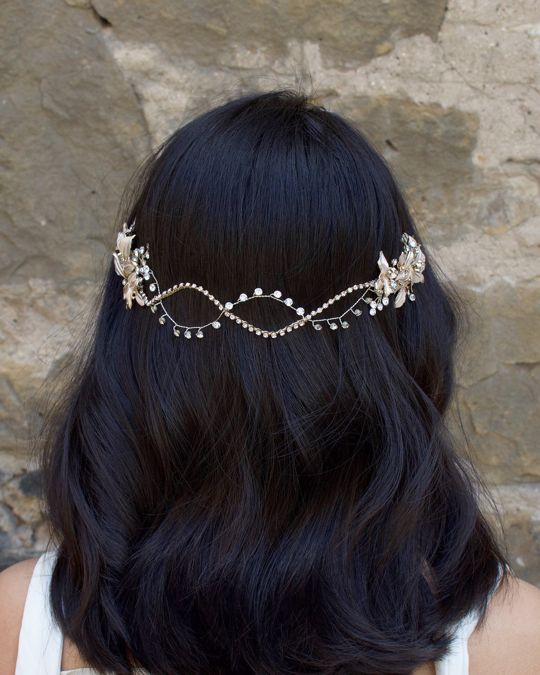 A Black haired model with her hair down wears a soft bridal vine at the back of her head. Stone wall background