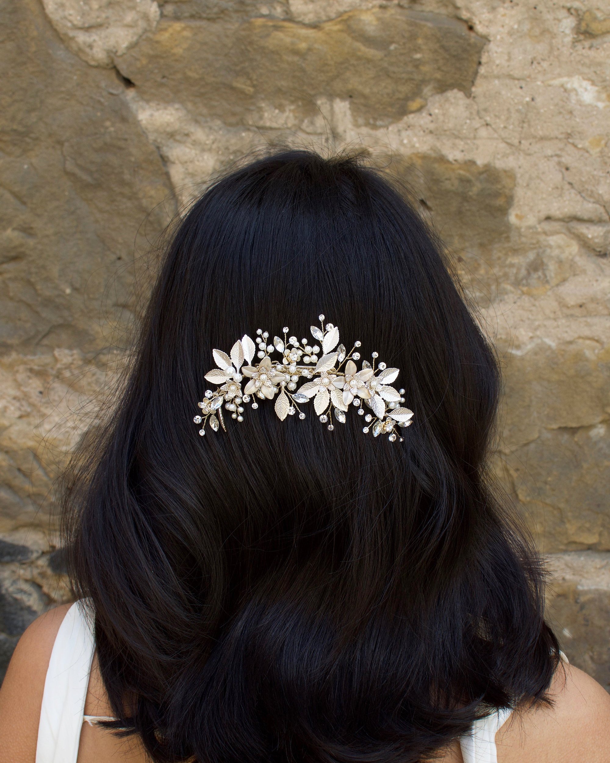 Dark haired model with long hair wears a side comb of pale gold leaves at the back of her head. With a stone wall background
