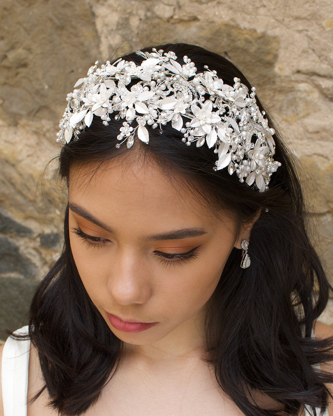 Black hair model wears a white leaves bridal headband that is wide with a stone wall behind her