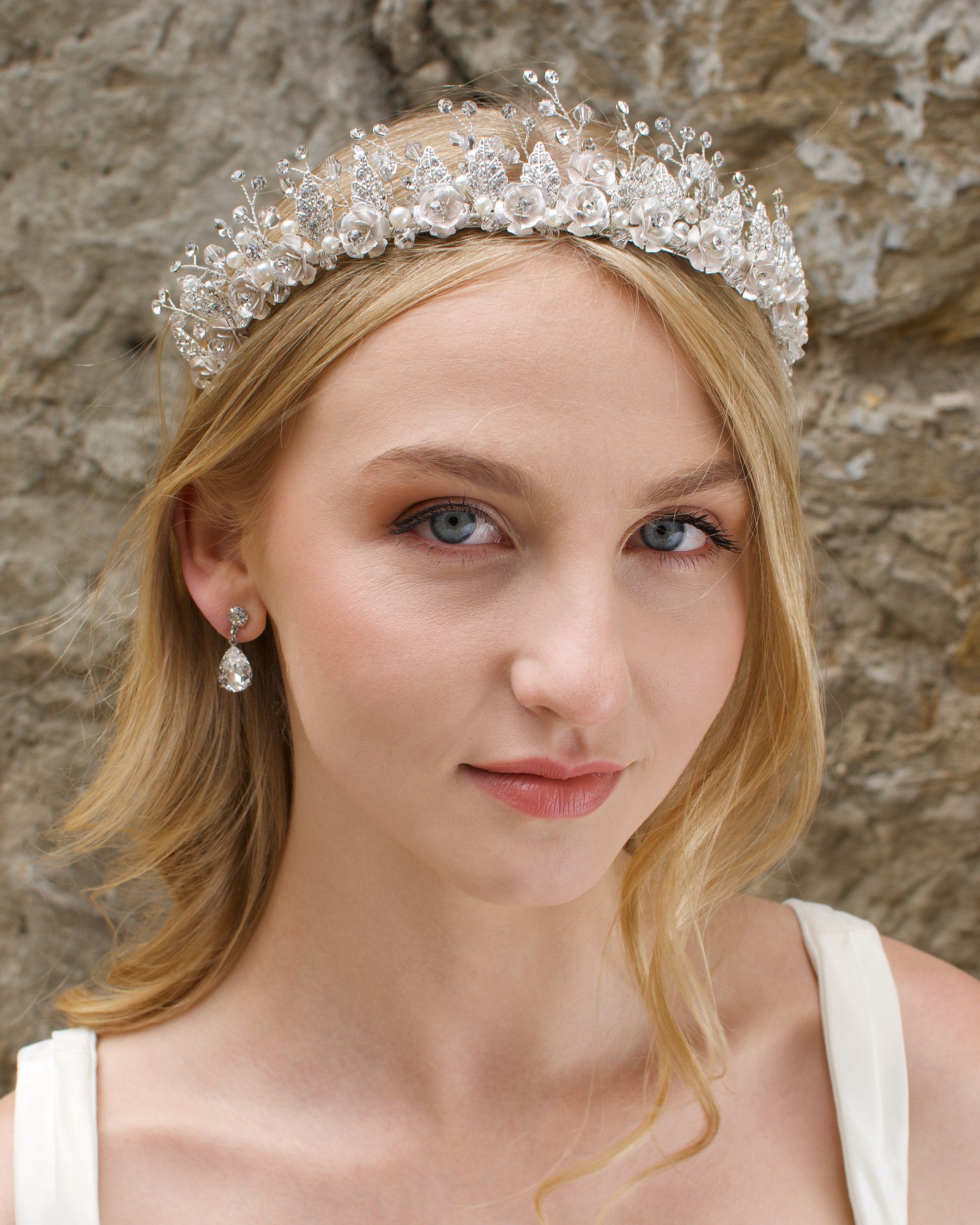 A Soft Silver Flowers and pearls wide tiara worn by a blonde bride with a stone wall background.