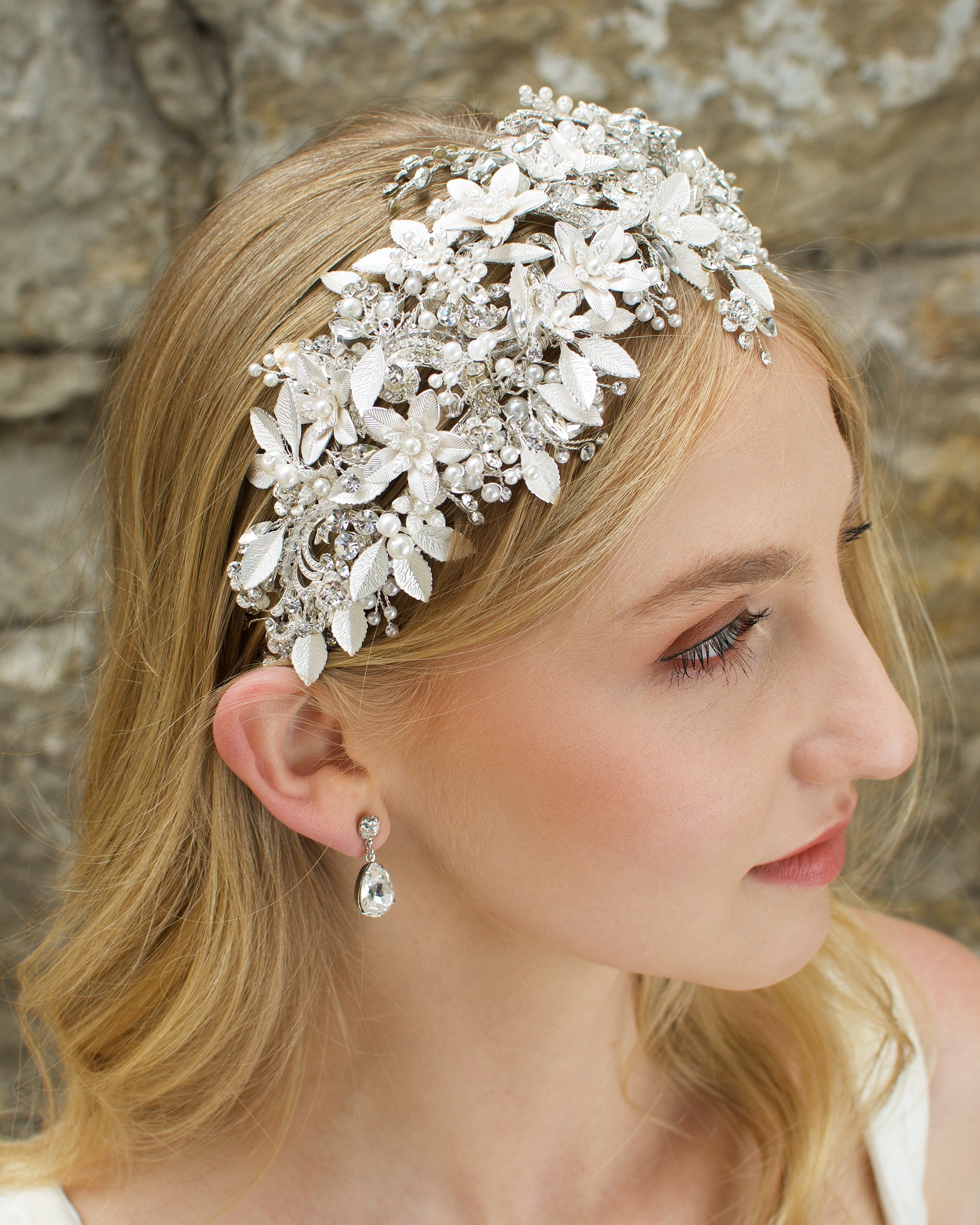 Blonde model wears a silver leaf headband thats wide she wears an earring and the background is a stone wall