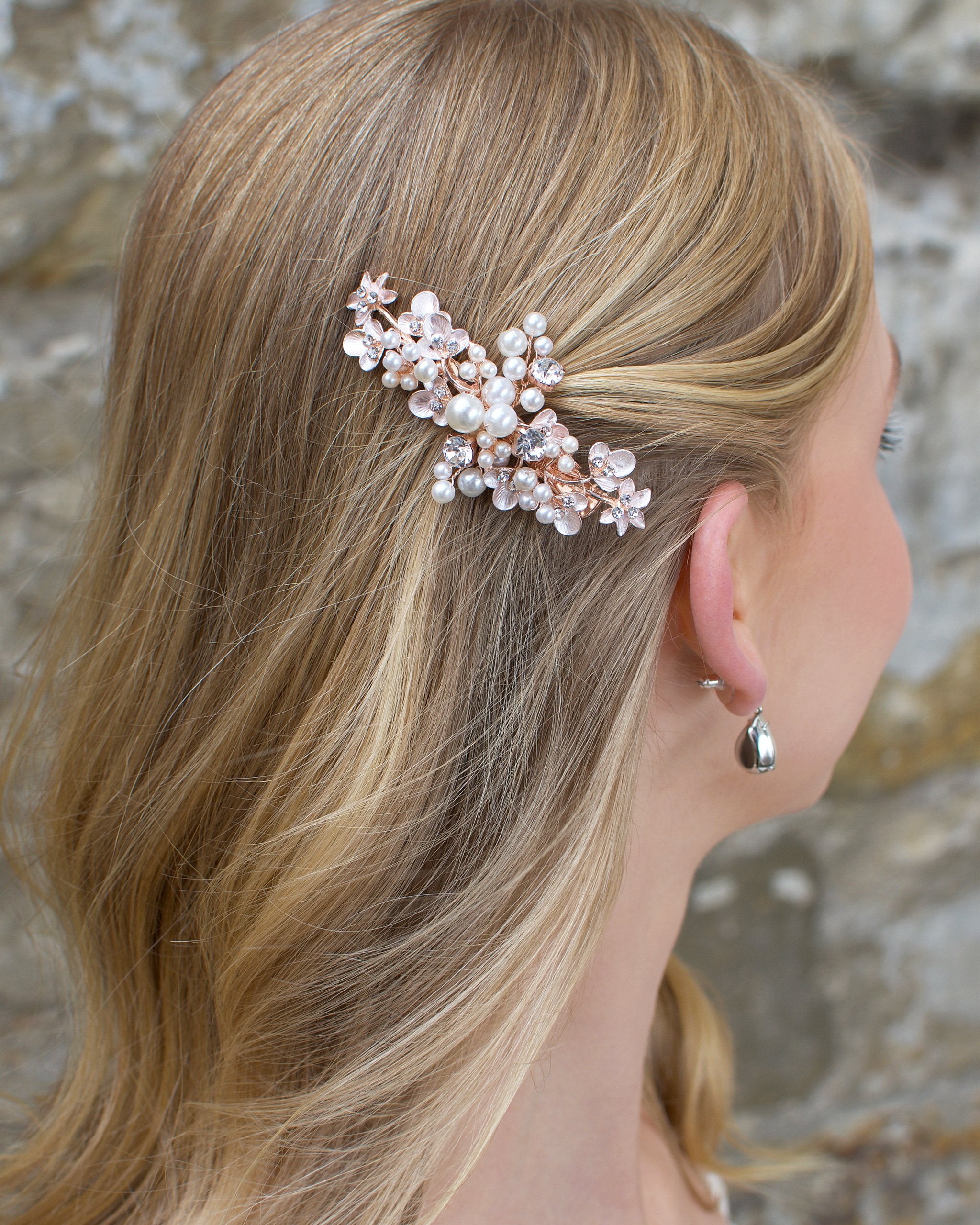 Blonde hair bride wears a pearl encrusted small side clip on rose gold with a stone wall behind.