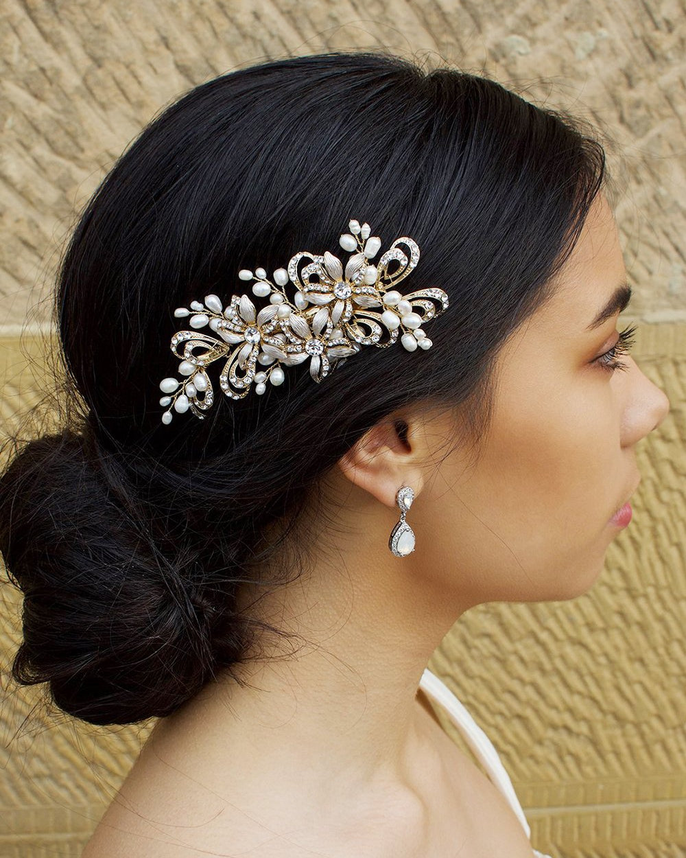 Model with dark hair wearing a small gold side comb in her hair. There is a stone wall behind her