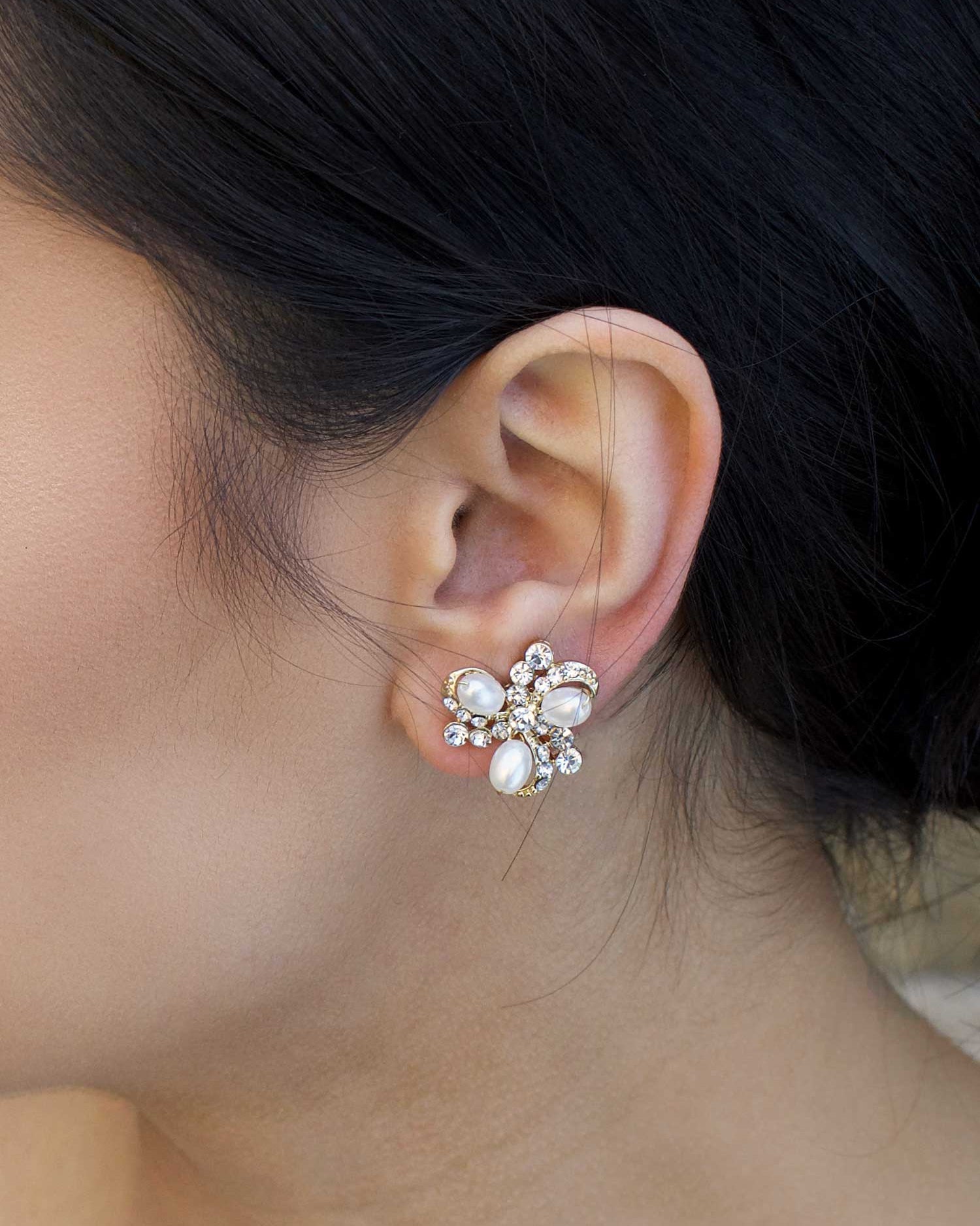 A close-up of a woman's ear wearing a pale gold hoop earring with three pearl studs, with tiny crystals embedded on the surface.