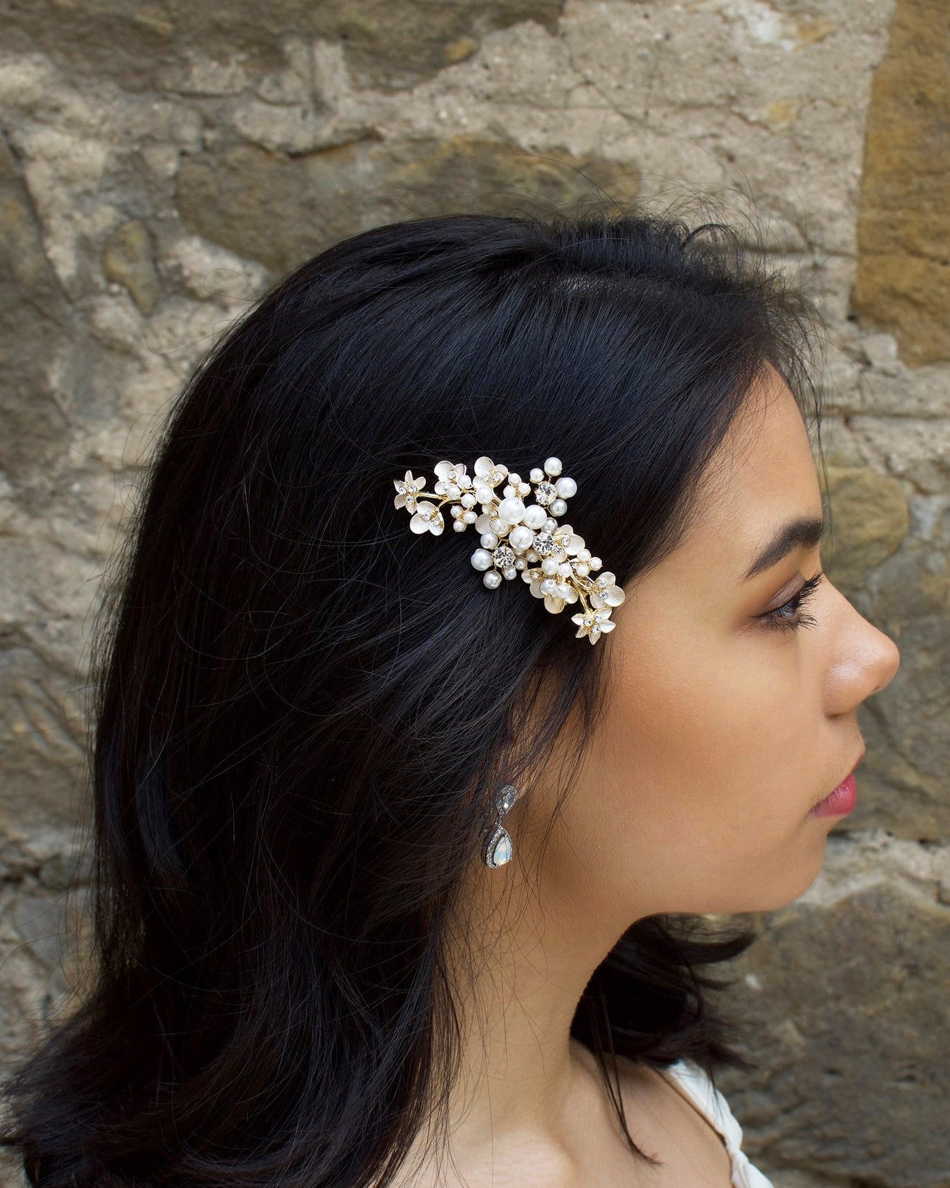 Our "Karina" Side Comb Pale Gold pearl clip worn by a model with dark hair against a stone wall