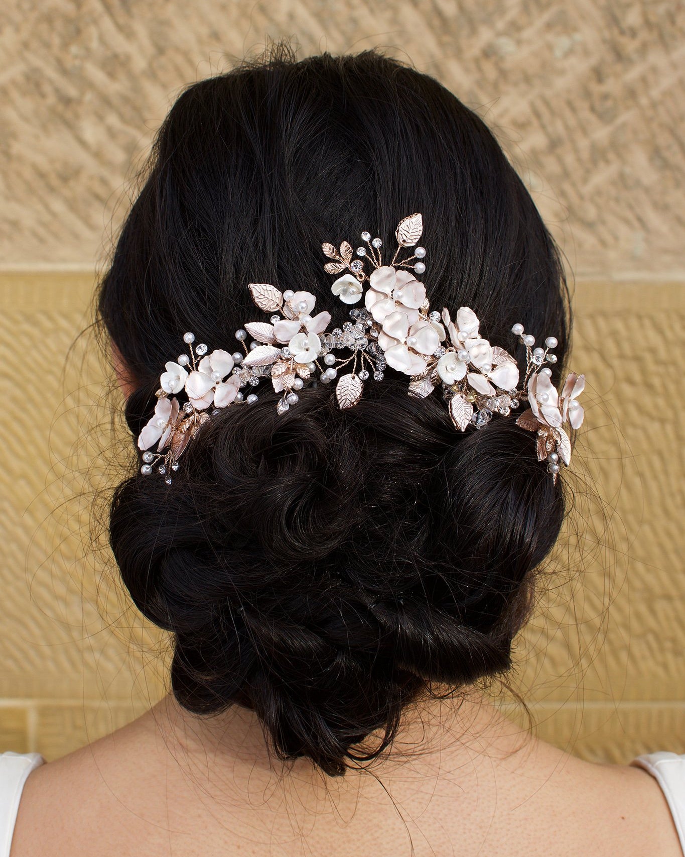 Bride with curling black hair wears a pale rose gold flowers headpiece on the back of her head. A stone wall is the background