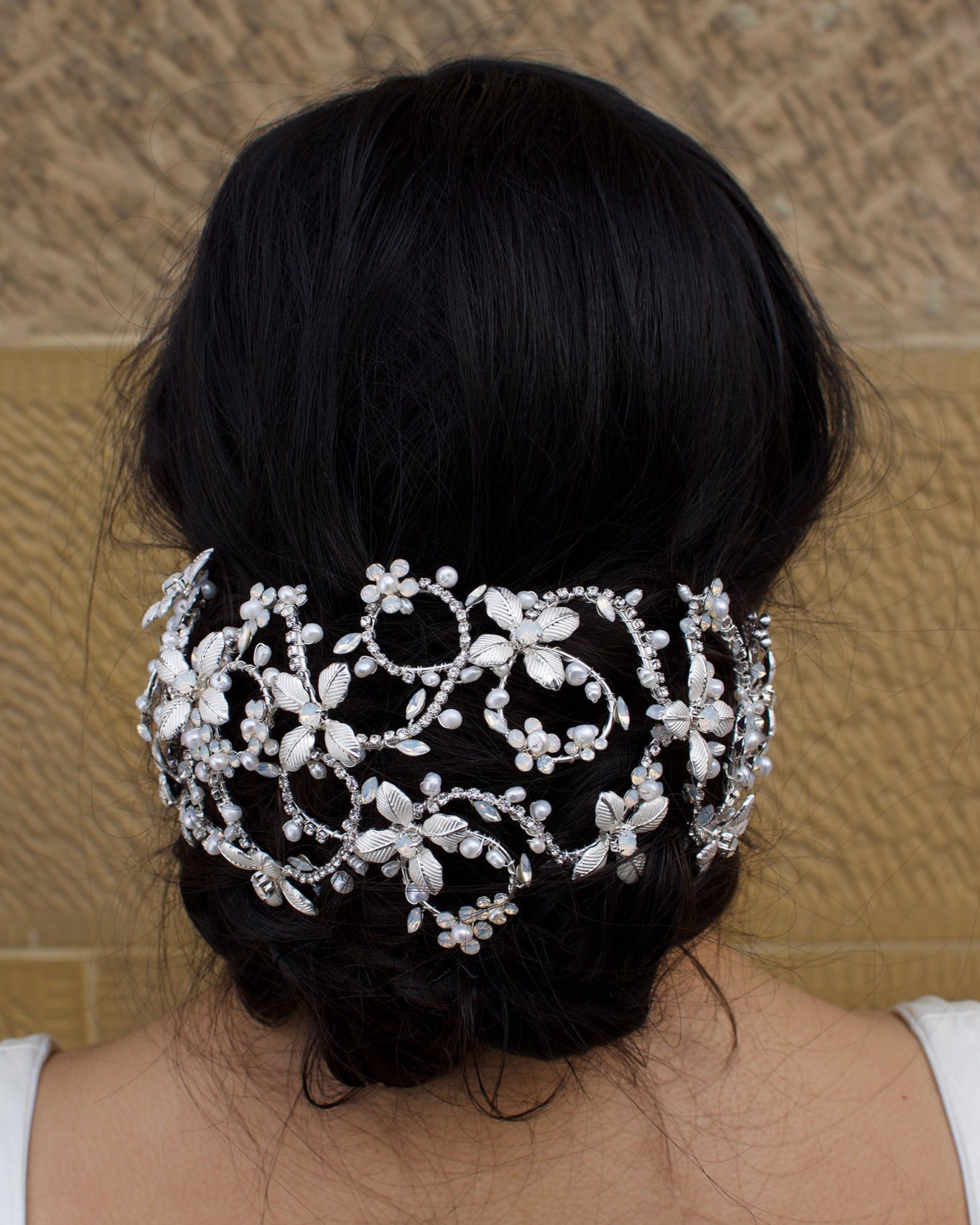 A black hair bridal model wears a wide silver hair cage on a bun of hair. There is a stone wall background