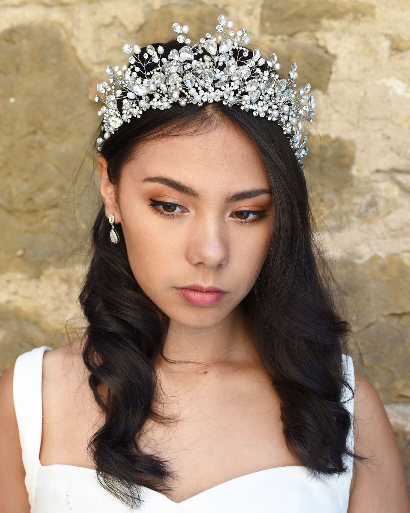 A model Bride wears a high Tiara of real pearls and crystals high on her head. She has dark hair and behind her is a stone wall.