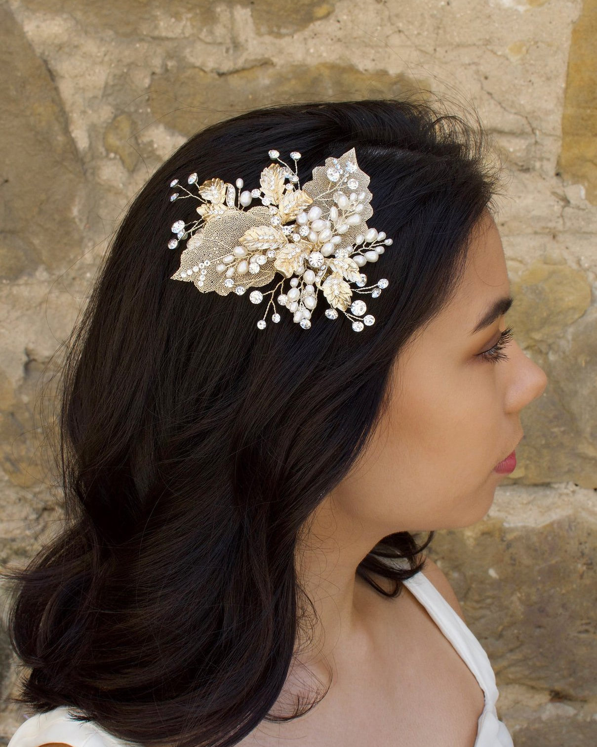 A bride with black hair wears a pale gold bridal hair clip with pearls in her dark hair in front of an old sandstone wall