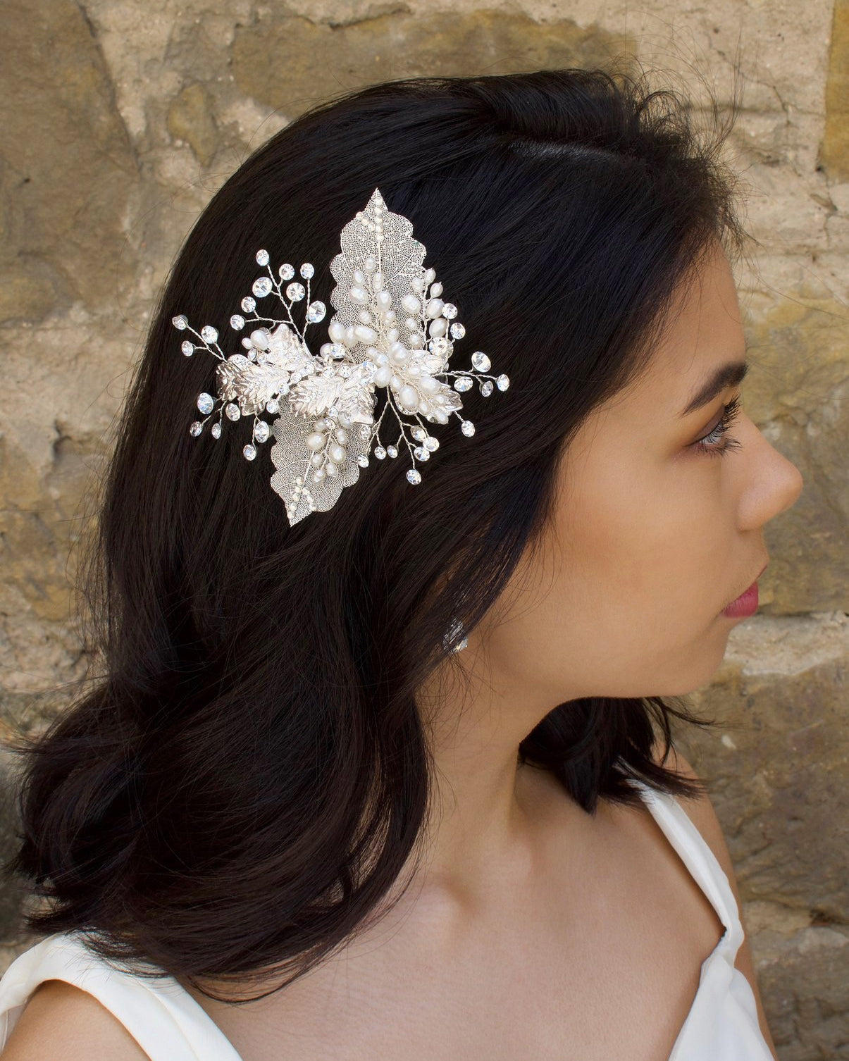 Dark hair bride wears a filigree leaf silver side comb with pearls in front of an old stone wall