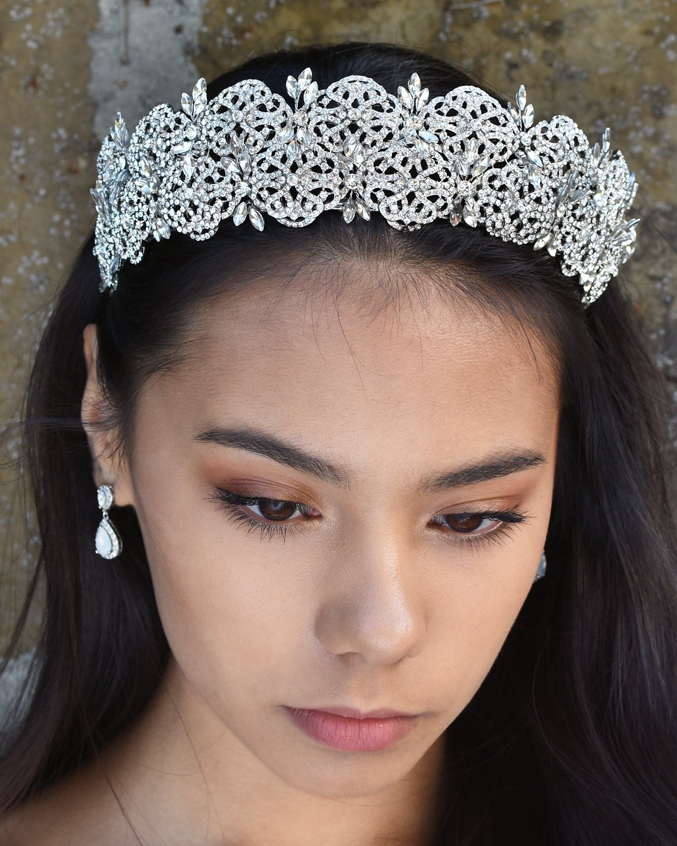 A wide crystal studded tiara is worn at the front of the head of a dark hair model.