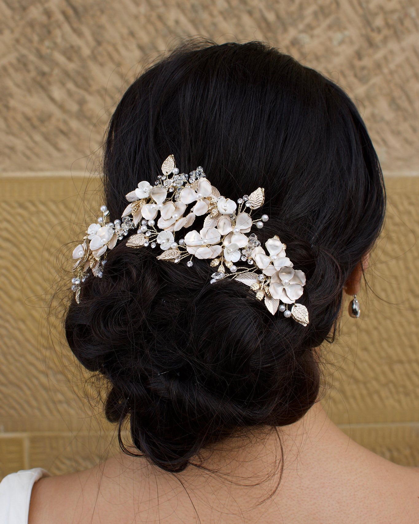 A dark haired bride wears a gold flowers headpiece at the back of her head with a stone wall background