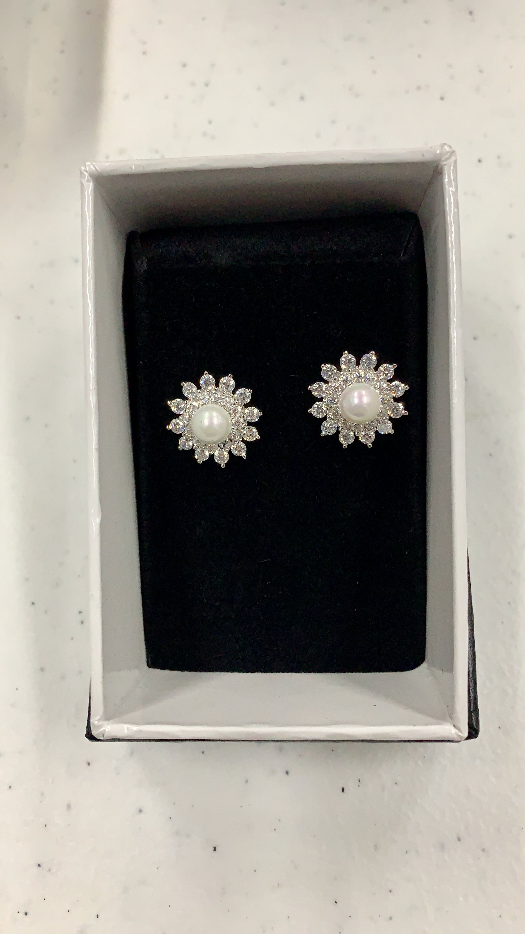 A pair of silver-toned flower-shaped stud earrings with white pearl centers and clear stones, presented in a white jewelry box with a black velvet insert.