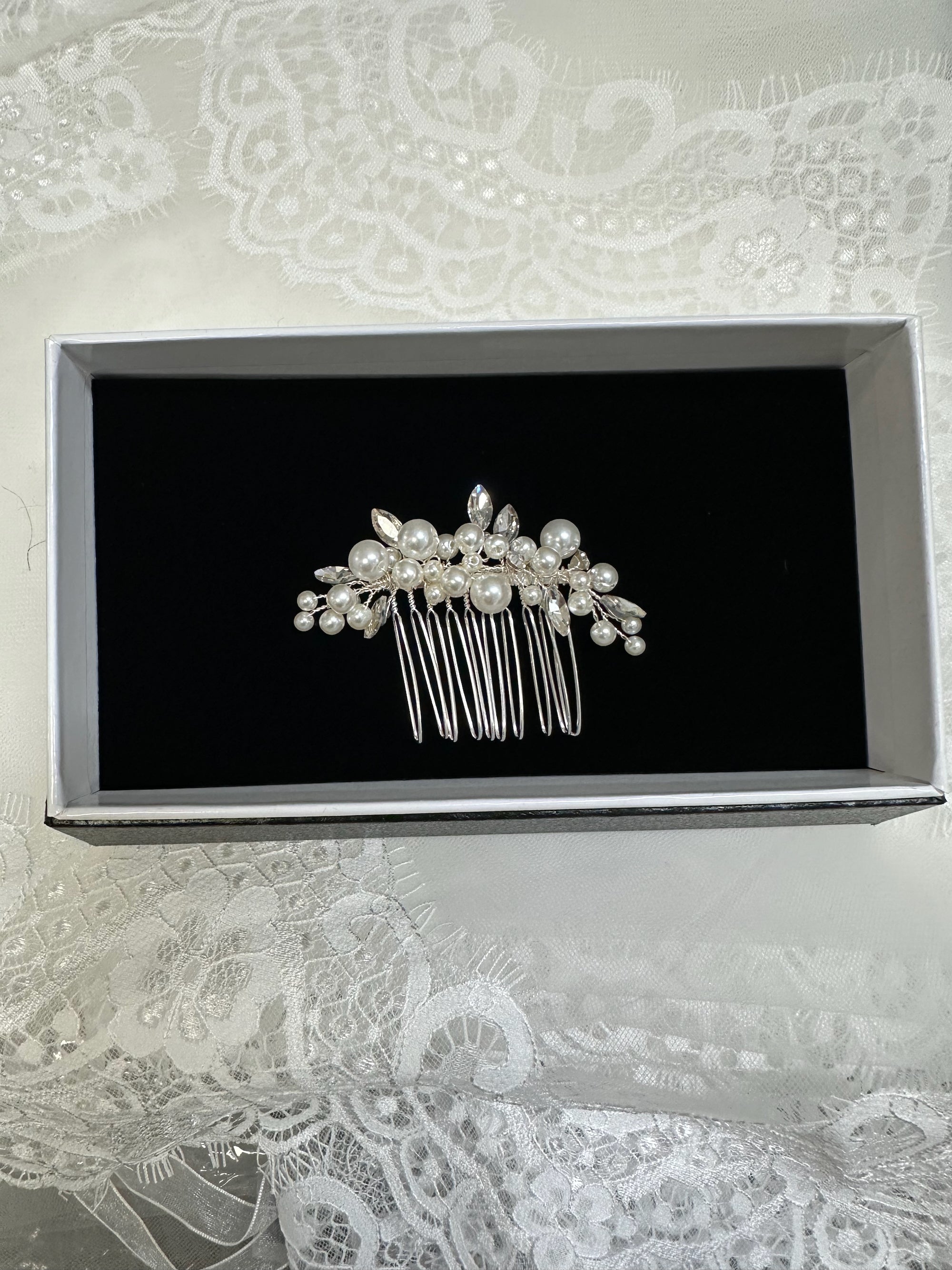 Silver hair comb with pearls on a black card inside a box, placed on lace fabric.