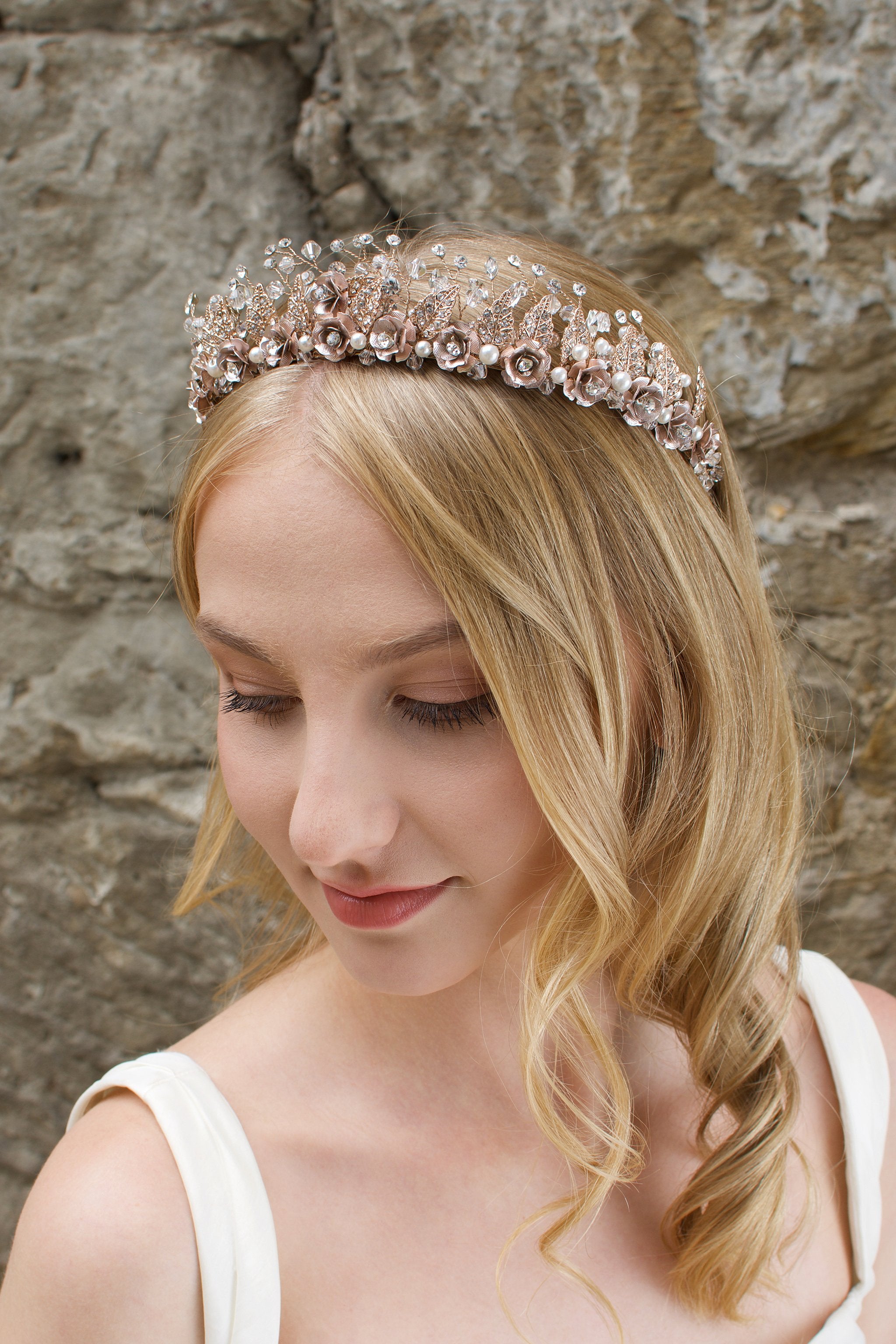 A Rose Gold Flowers and pearls wide tiara worn by a blonde bride with a stone wall background.