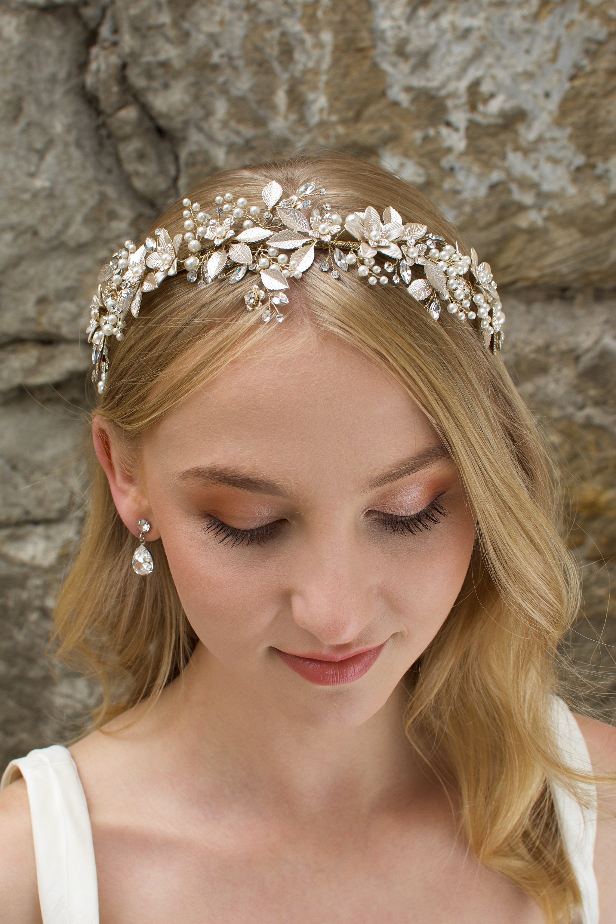 A model Bride with blonde hair wears a gold bridal crown at the front of her head with a stone wall behind