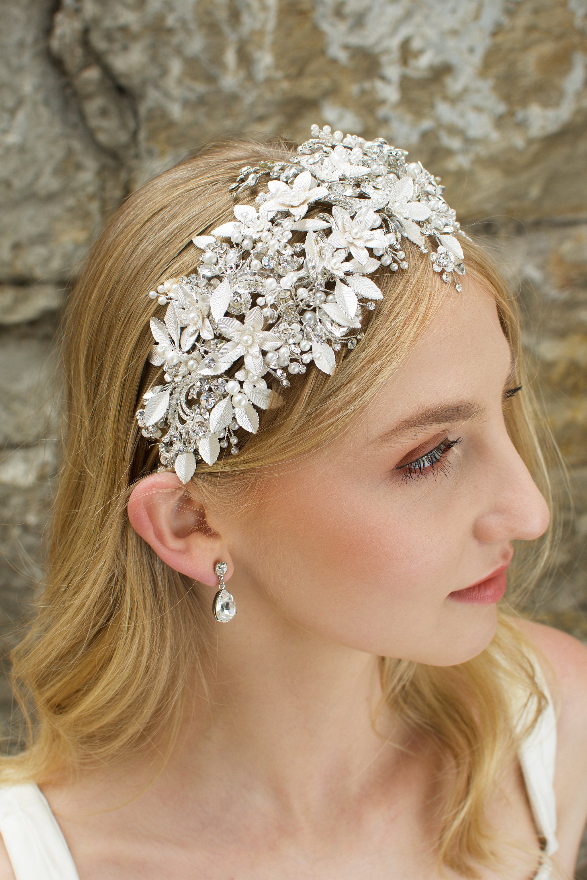 Blonde model wears a silver leaf headband thats wide she wears an earring and the background is a stone wall