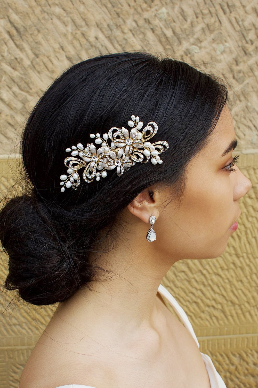 Model with dark hair wearing a small gold side comb in her hair. There is a stone wall behind her