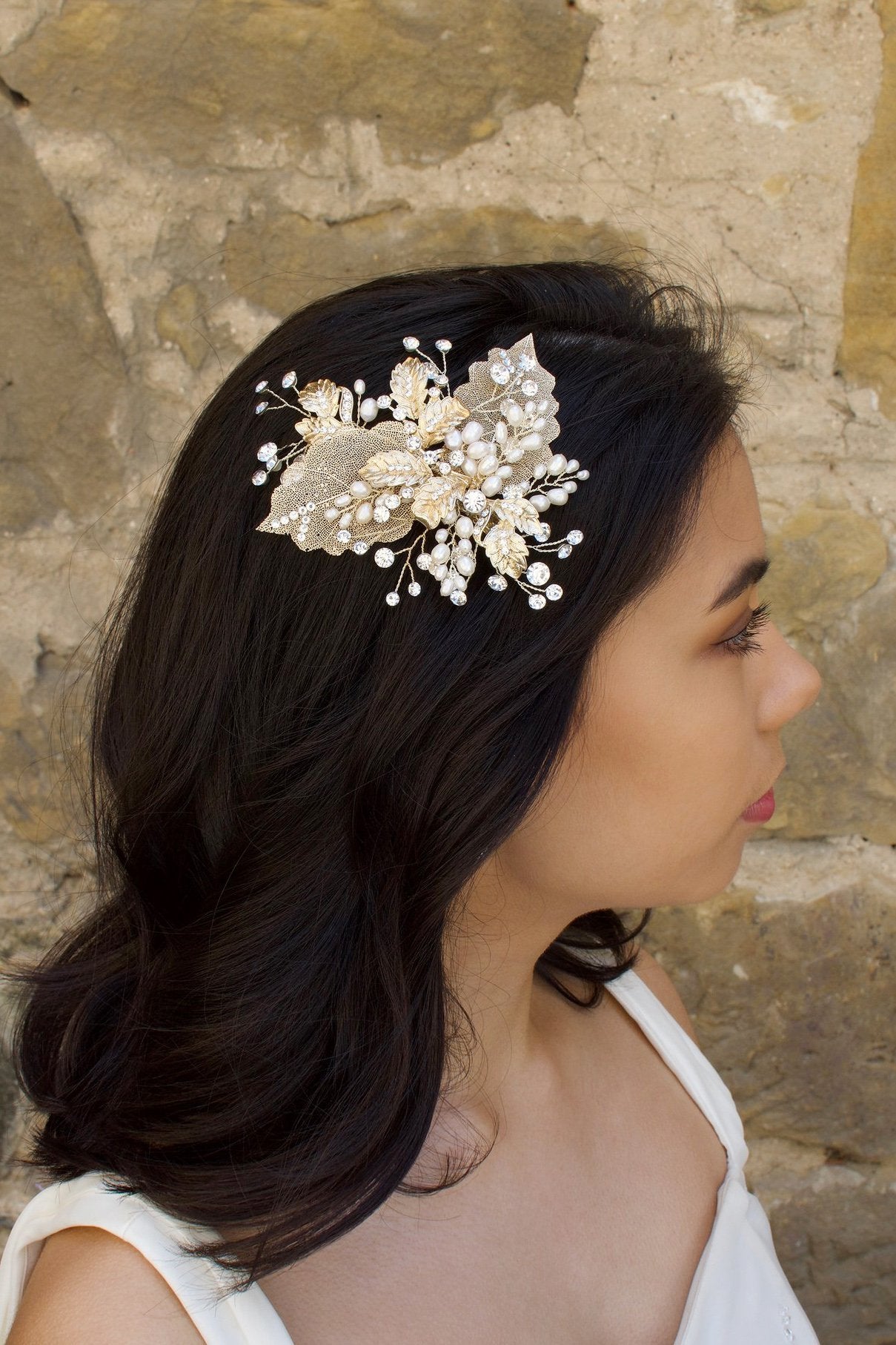 A bride with black hair wears a pale gold bridal hair clip with pearls in her dark hair in front of an old sandstone wall