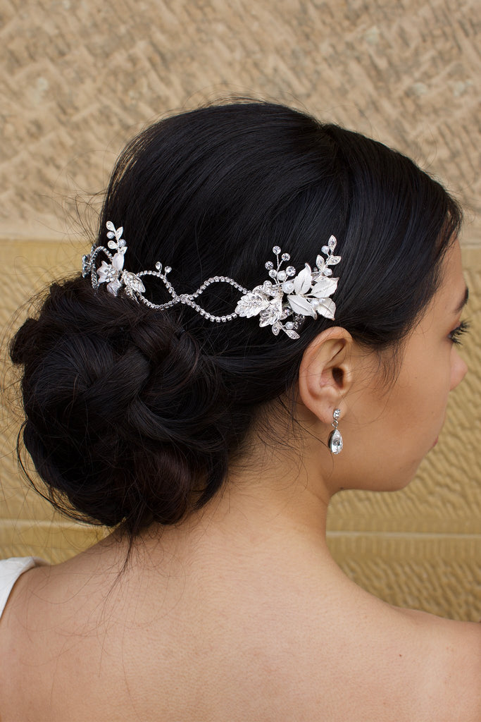 A dark haired model with curly hair wears a silver vine at the back of her head with a stone wall behind